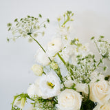 A close-up of a white floral bouquet featuring roses, ranunculus, and delicate green foliage against a soft light gray background available for pick-up and delivery in Columbus, Ohio