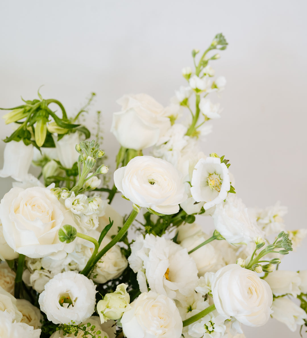 A close-up of a white floral bouquet featuring roses, ranunculus, and green foliage against a soft light gray background.