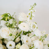 A close-up of a white floral bouquet featuring roses, ranunculus, and green foliage against a soft light gray background.