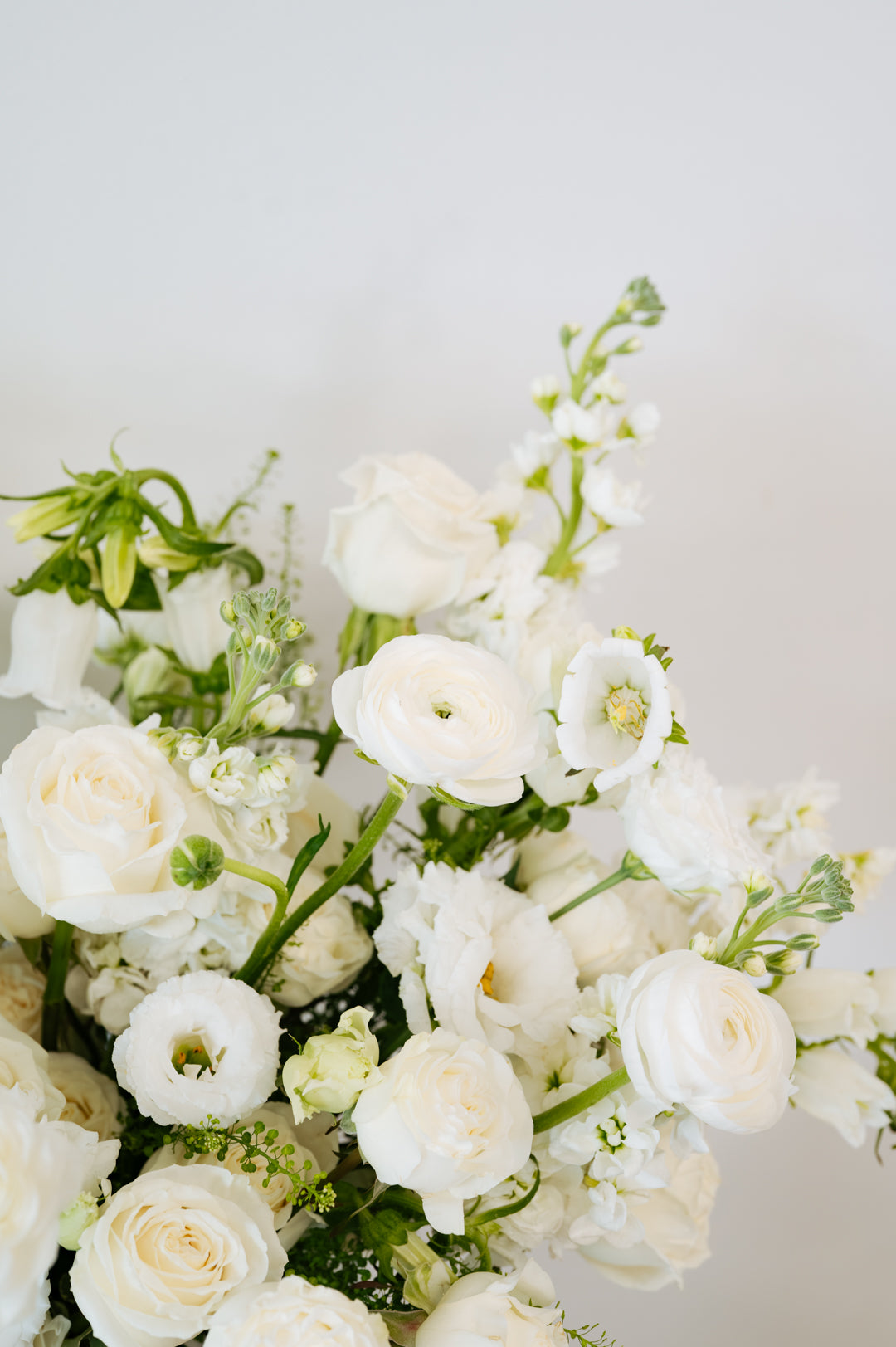 A close-up of a white floral bouquet featuring roses, ranunculus, and green foliage against a soft light gray background.