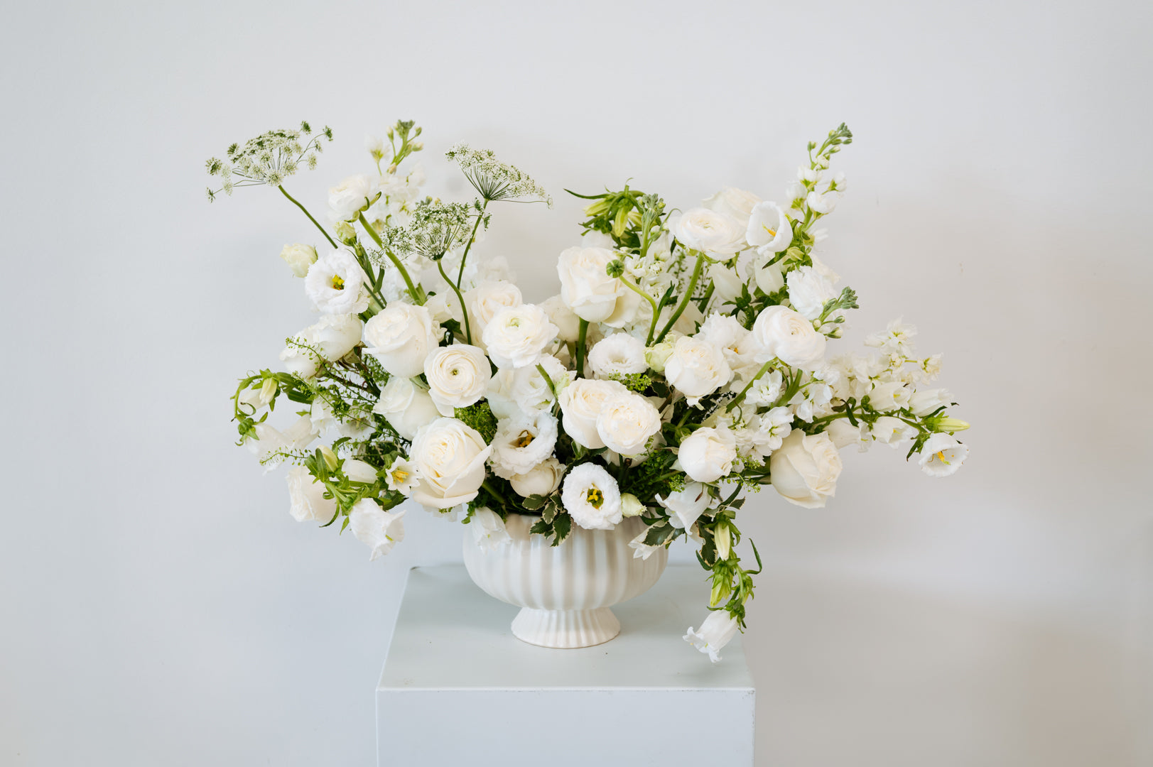 A white ceramic urn-shaped vase filled with a lush arrangement of white flowers and green foliage, displayed on a white pedestal against a light gray background available for local pick-up and delivery in Columbus, Ohio