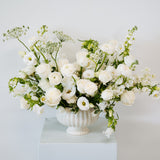 A white ceramic urn-shaped vase filled with a lush arrangement of white flowers and green foliage, displayed on a white pedestal against a light gray background available for local pick-up and delivery in Columbus, Ohio
