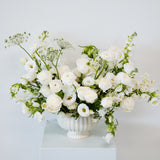 A white ceramic urn-shaped vase filled with a lush bouquet of white roses, white baby's breath, and green foliage, displayed on a white pedestal against a white background available for local pick-up and delivery in Columbus, Ohio