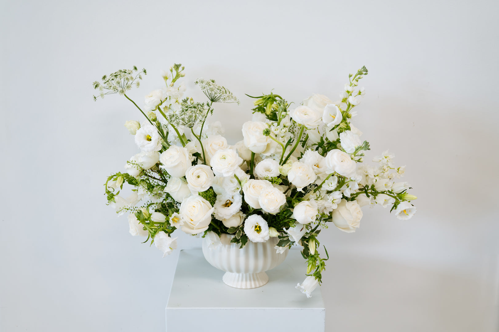 A white ceramic urn-shaped vase filled with a lush bouquet of white roses, white baby's breath, and green foliage, displayed on a white pedestal against a white background available for local pick-up and delivery in Columbus, Ohio