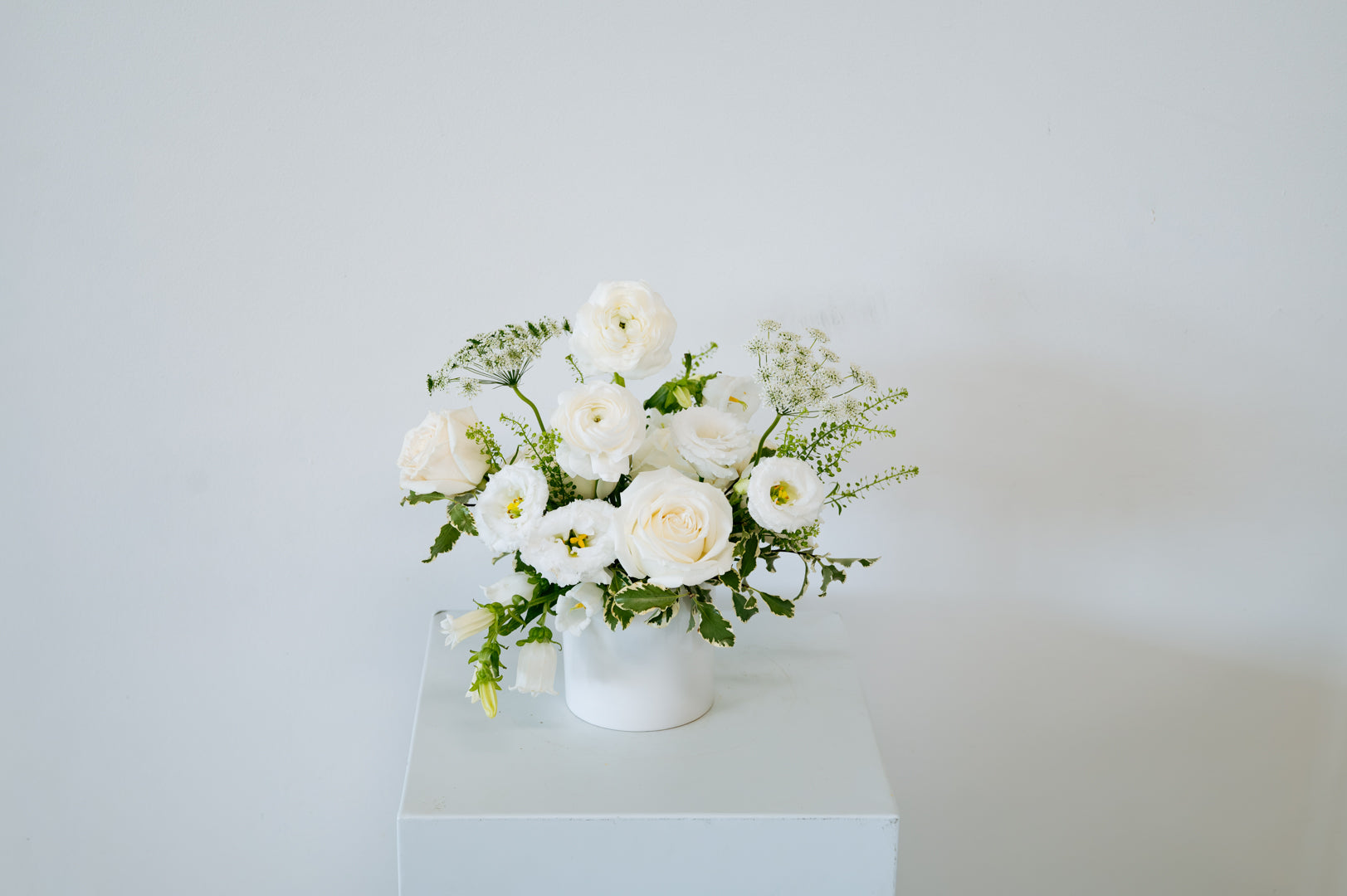 A white ceramic vase filled with a fresh floral arrangement of white roses, white ranunculus, and green foliage, displayed on a white pedestal against a plain white wall available for local pick-up and delivery in Columbus, Ohio