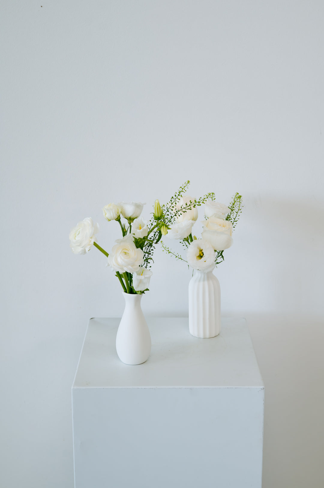 Two white ceramic vases with white flowers and green stems, displayed on a white pedestal against a light gray wall available for pick-up and local delivery in Columbus, Ohio