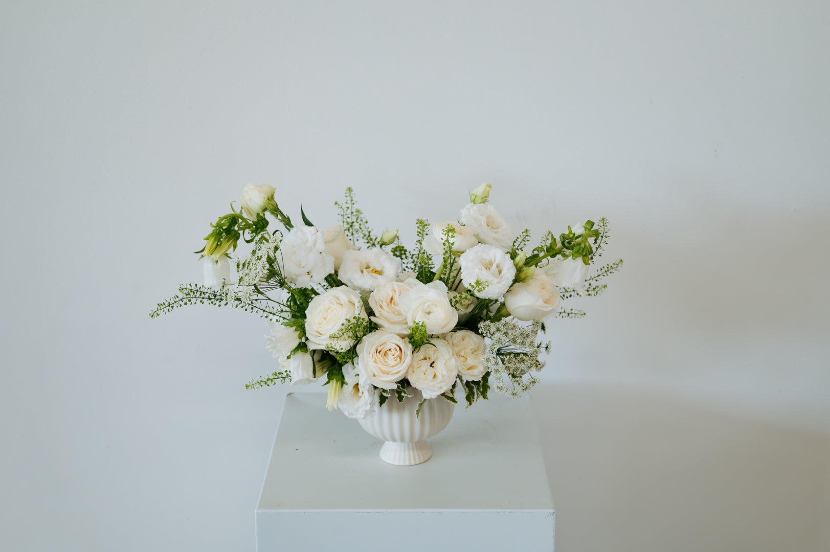 A white floral arrangement featuring white roses, peonies, and green foliage, presented in a white ribbed ceramic urn on a white pedestal against a plain white background available for local delivery and pick-up in Columbus, Ohio