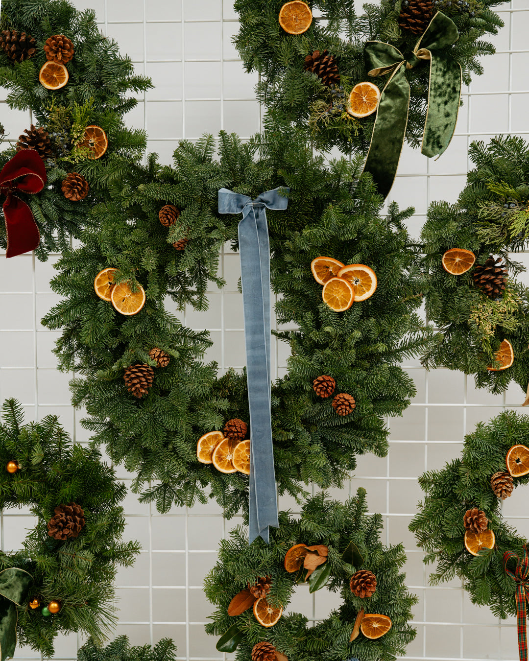 Evergreen wreaths and garlands hang on a white tiled wall, decorated with dried orange slices, pinecones, and ribbons for a warm festive feel.